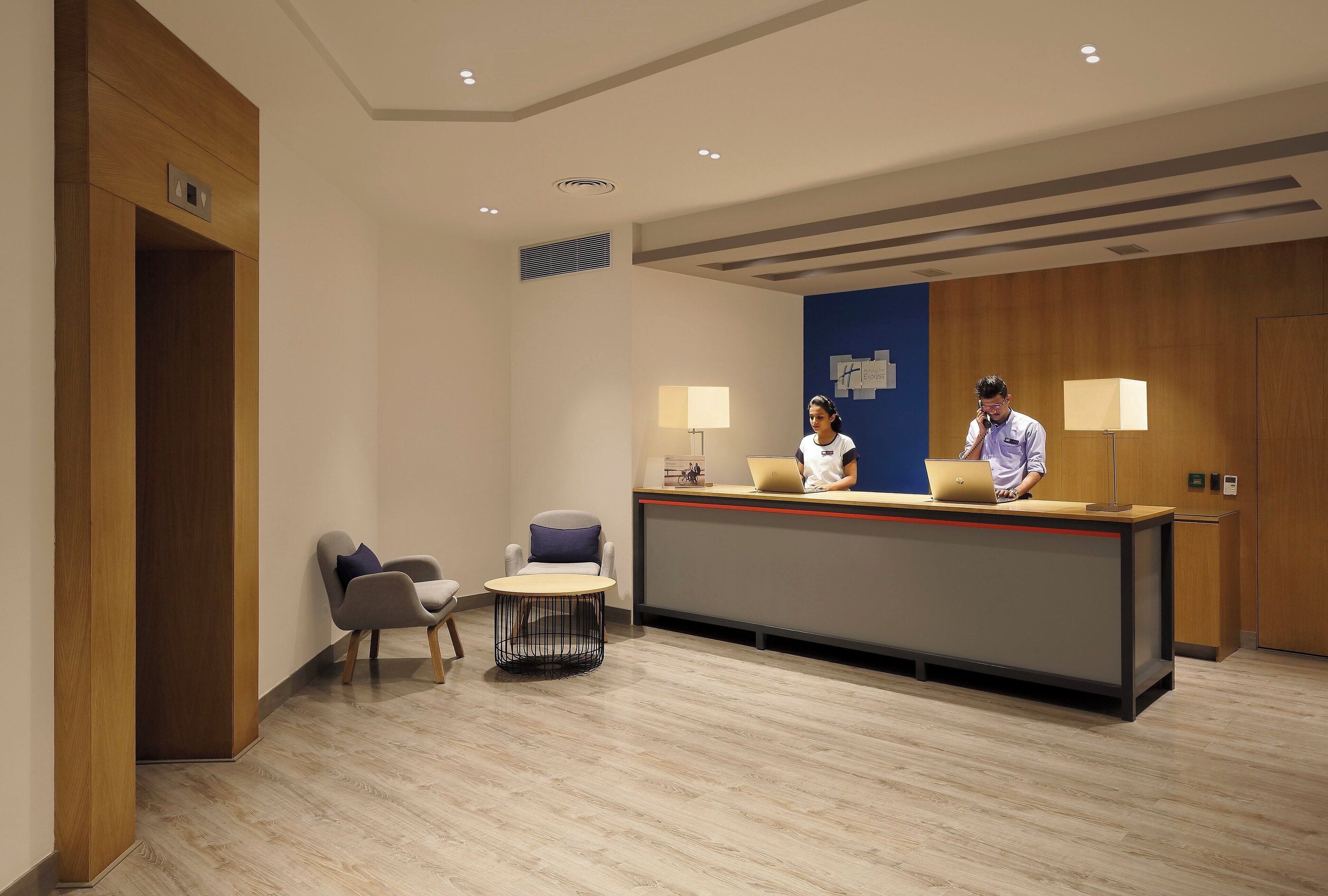 Un espace d’accueil d’hôtel avec un comptoir de réception en bois, deux chaises, une petite table et un mur bleu orné d’une enseigne.