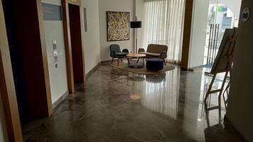 A modern lobby with a polished marble floor, a seating area with a round table and chairs, a large abstract painting, and an elevator.