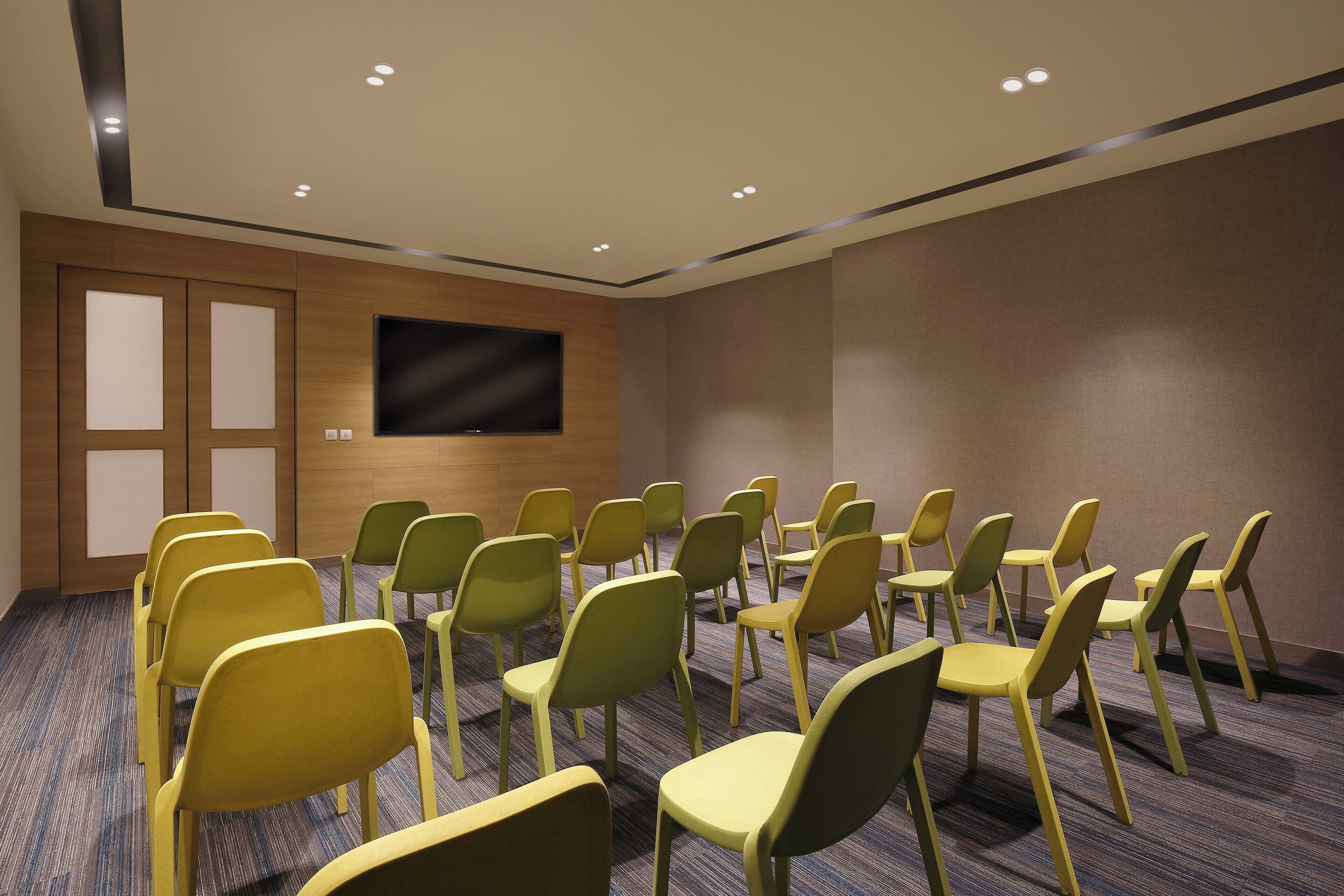 A conference room with rows of chairs, a large flat-screen TV on the wall, and a wooden door.