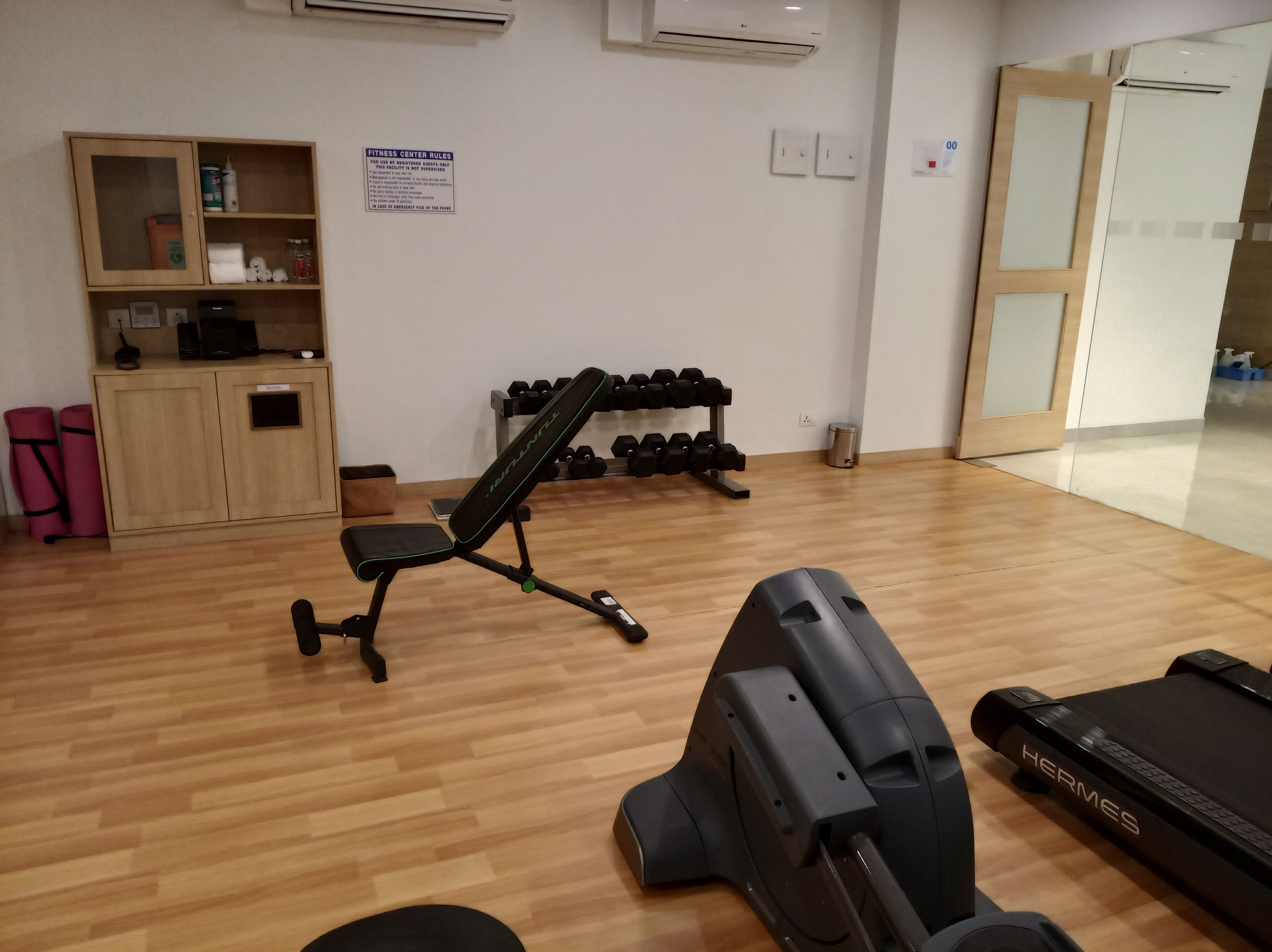 A fitness center with a bench, dumbbells, a treadmill, and a cabinet with towels and a water bottle.