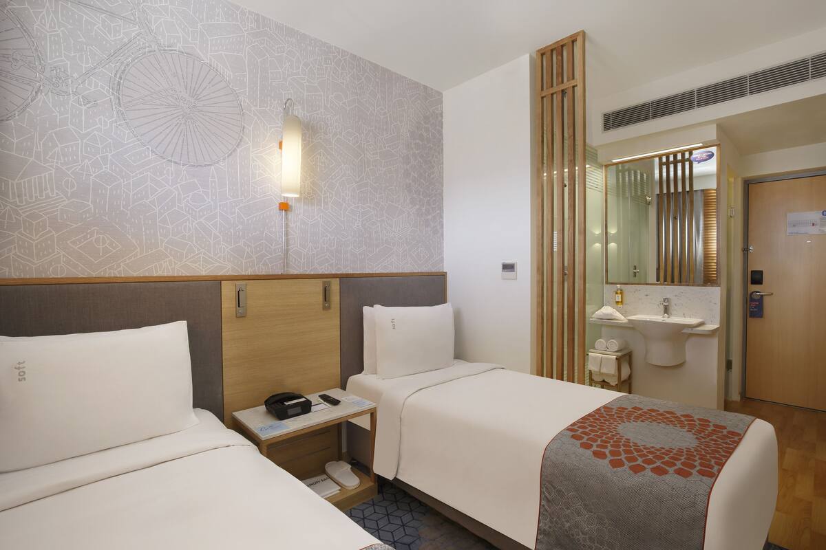 Ein Hotelzimmer mit zwei Betten, einem Nachttisch mit Telefon und Lampe, einem Badezimmer mit Waschbecken und Spiegel sowie einer Wand mit Muster.