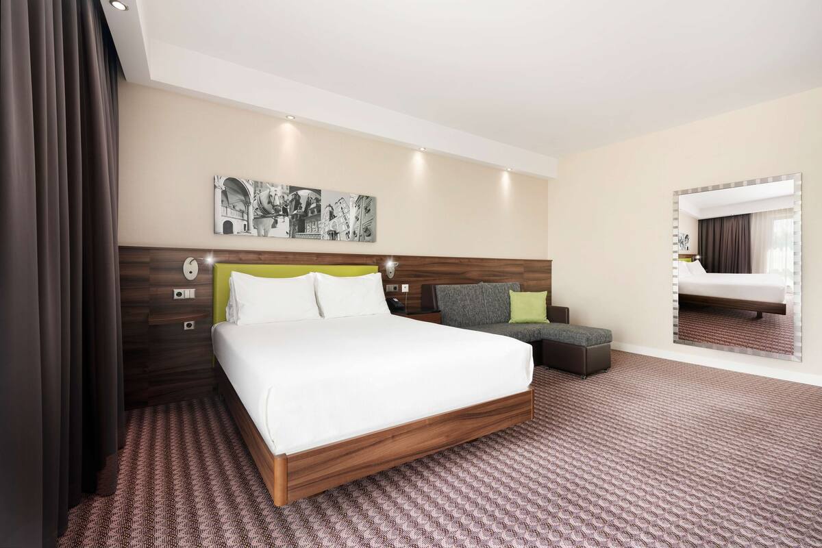 A modern hotel room with a large bed, a small sofa, a mirror, and a wall-mounted artwork.