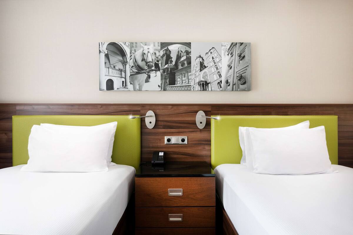 A hotel room with two beds, a wooden headboard, a nightstand with a phone, and a wall-mounted artwork depicting a horse and buildings.