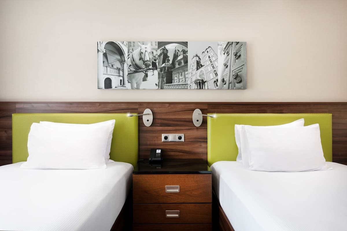 Quarto de hotel com duas camas, cabeceira de madeira, mesa de cabeceira com telefone e uma obra de arte na parede retratando um cavalo e edifícios.