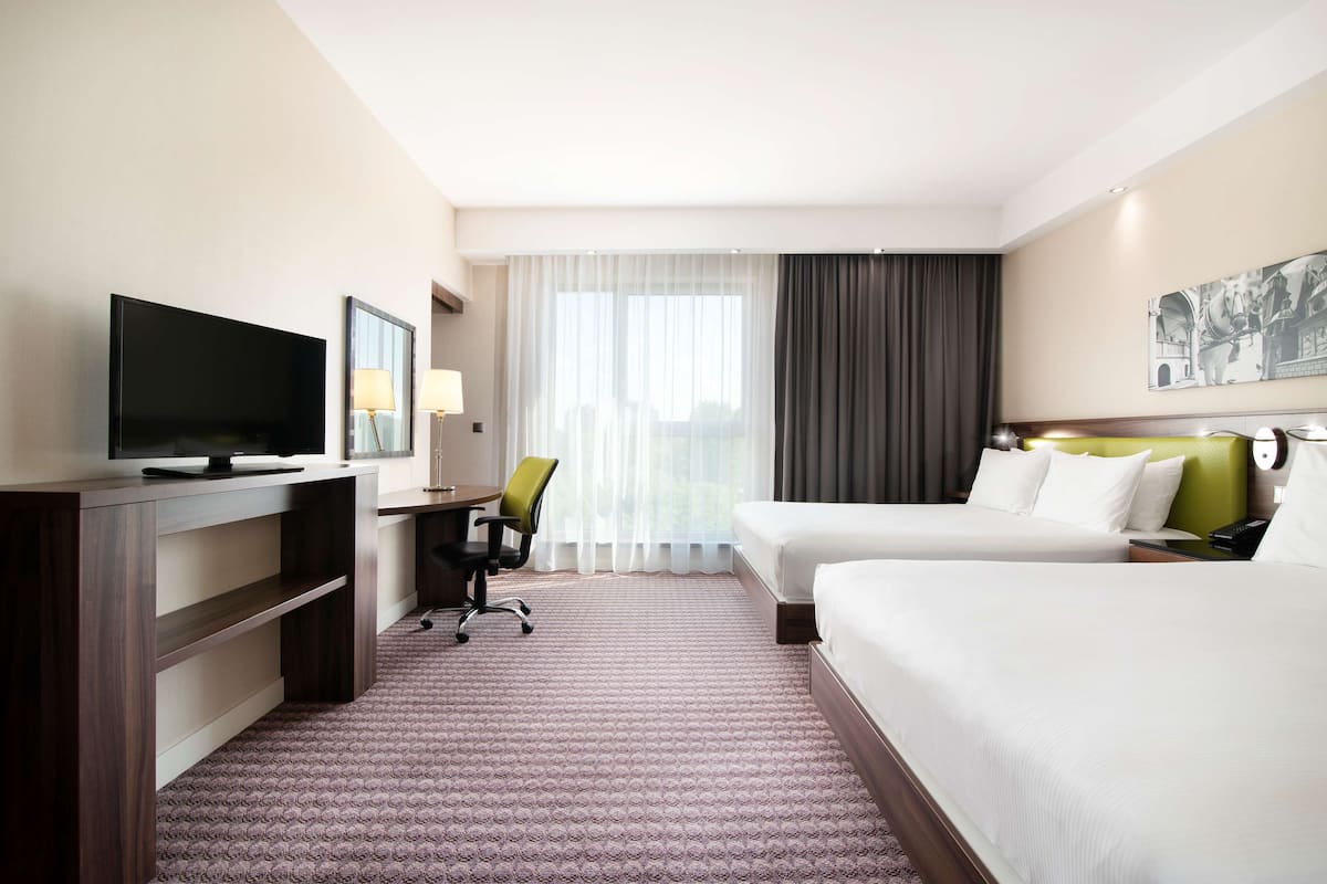 A hotel room with two beds, a desk with a chair, a television, and a large window with curtains.