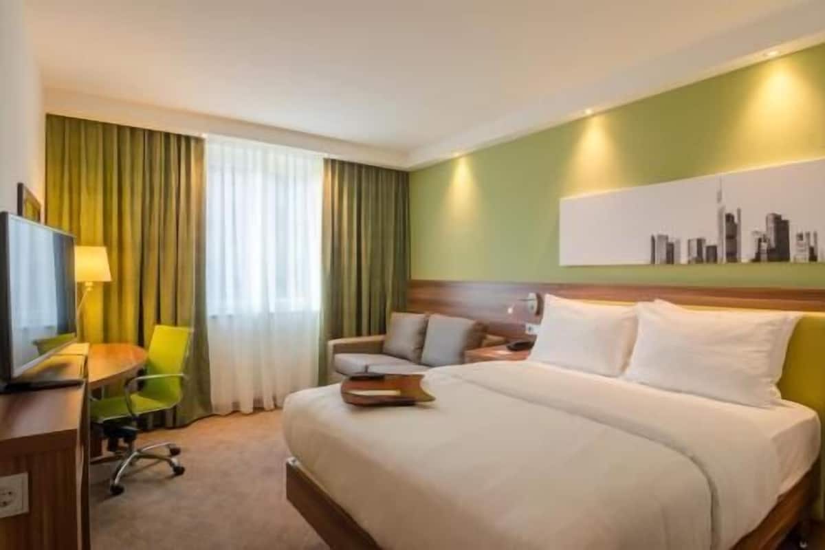 Habitación de hotel con cama, escritorio con silla, televisor y un cuadro de paisaje urbano en la pared.