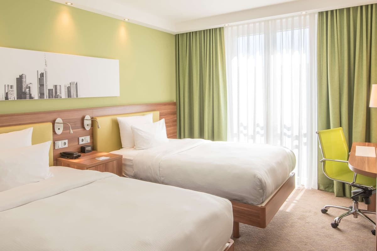 Habitación de hotel con dos camas, un escritorio con silla, un televisor y un cuadro de la ciudad en la pared.