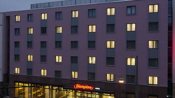 Ein mehrstöckiges Hotel mit schlichter Fassade, großen Fenstern und einem leuchtenden Neonzeichen mit dem Schriftzug 'Hampton'.