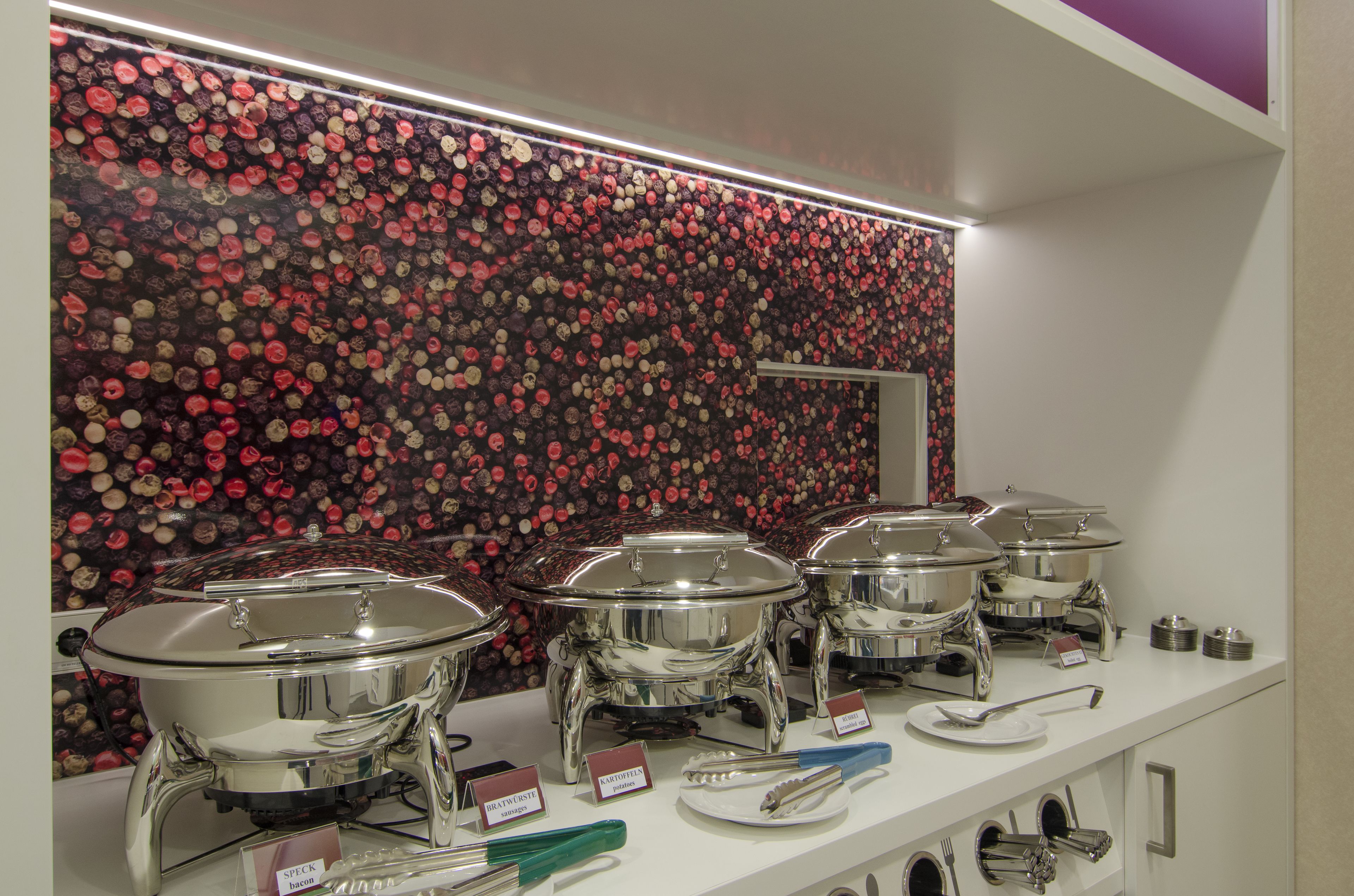 Un espace buffet avec plusieurs plats de service en acier inoxydable disposés sur un comptoir blanc, un mur décoré d’un motif d’objets rouges et noirs, ainsi que divers ustensiles et assiettes de service.
