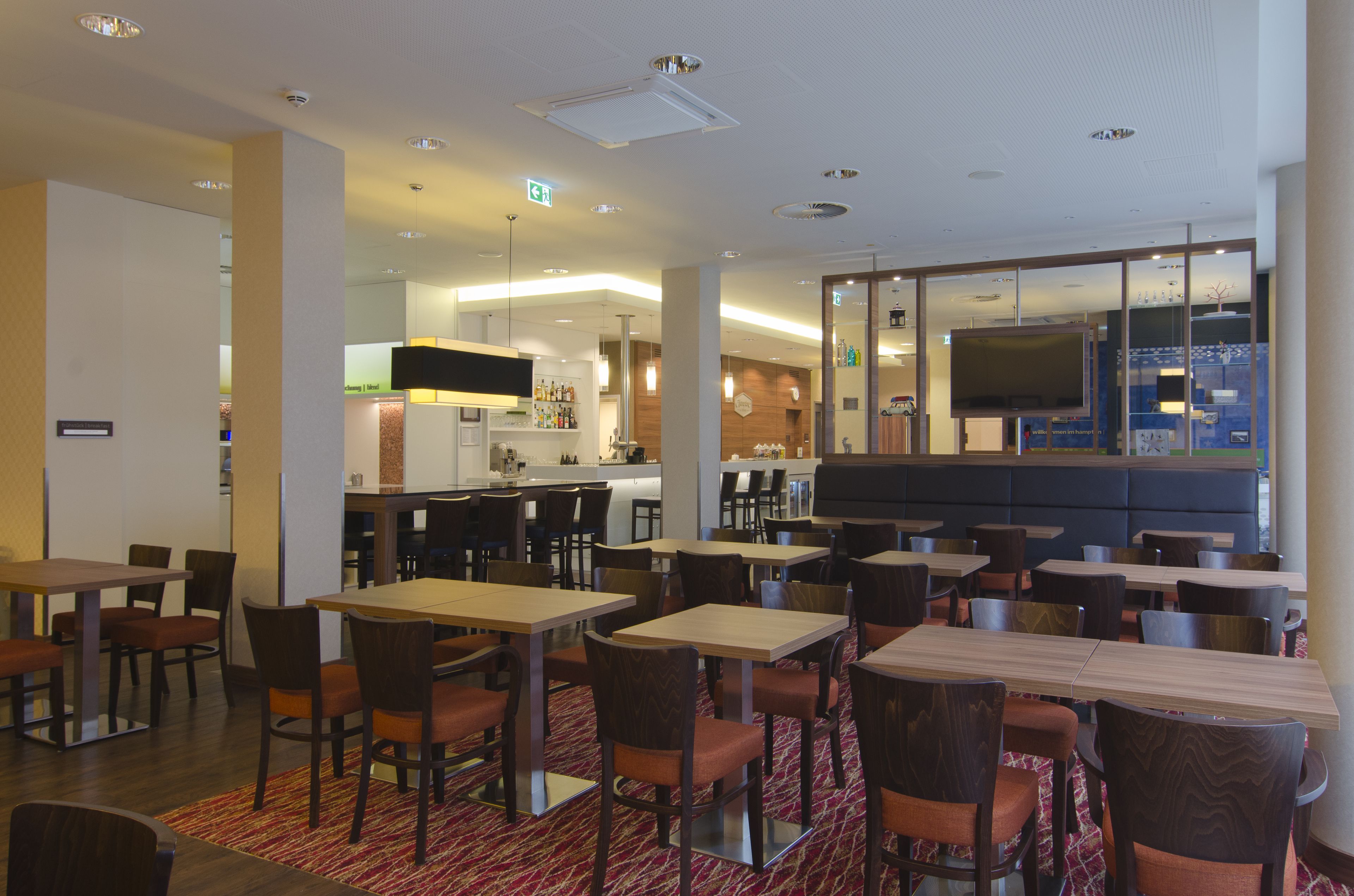 A hotel dining area with multiple tables and chairs, a kitchen in the background, and a large TV screen.
