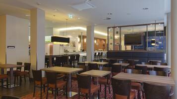 A hotel dining area with multiple tables and chairs, a kitchen in the background, and a large TV screen.