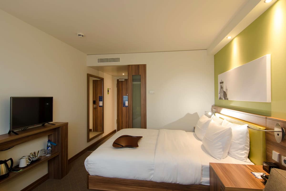 Una habitación de hotel con cama, mesita de noche, televisor y repisa de madera con artículos de aseo.