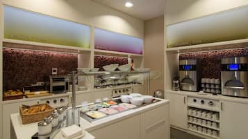Zona de desayuno tipo buffet con diversas opciones de comida, una estación de café y una selección de tazas y cuencos.