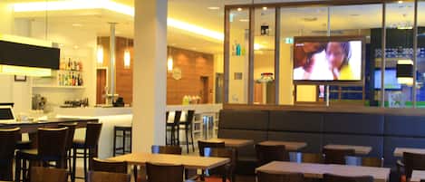 Un restaurante con varias mesas y sillas, una zona de bar y una pantalla de televisión mostrando un programa.