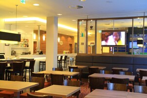 A restaurant with multiple tables and chairs, a bar area, and a television screen displaying a program.