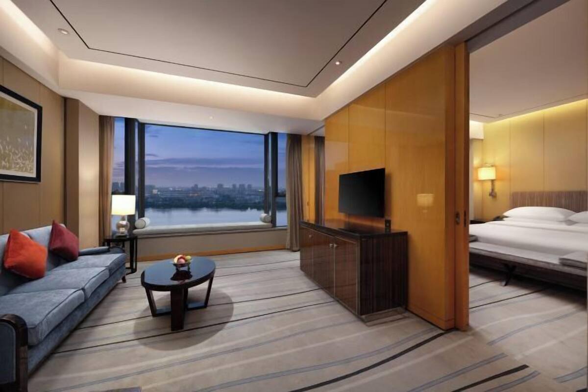 A hotel room with a large window, a sofa, a TV, a bed, and a small table.