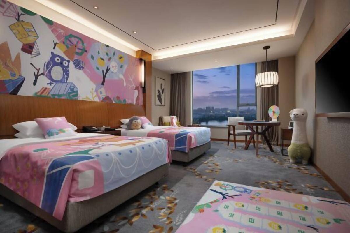 A hotel room with two beds, a large window with a city view, a small table, and a decorative wall mural.