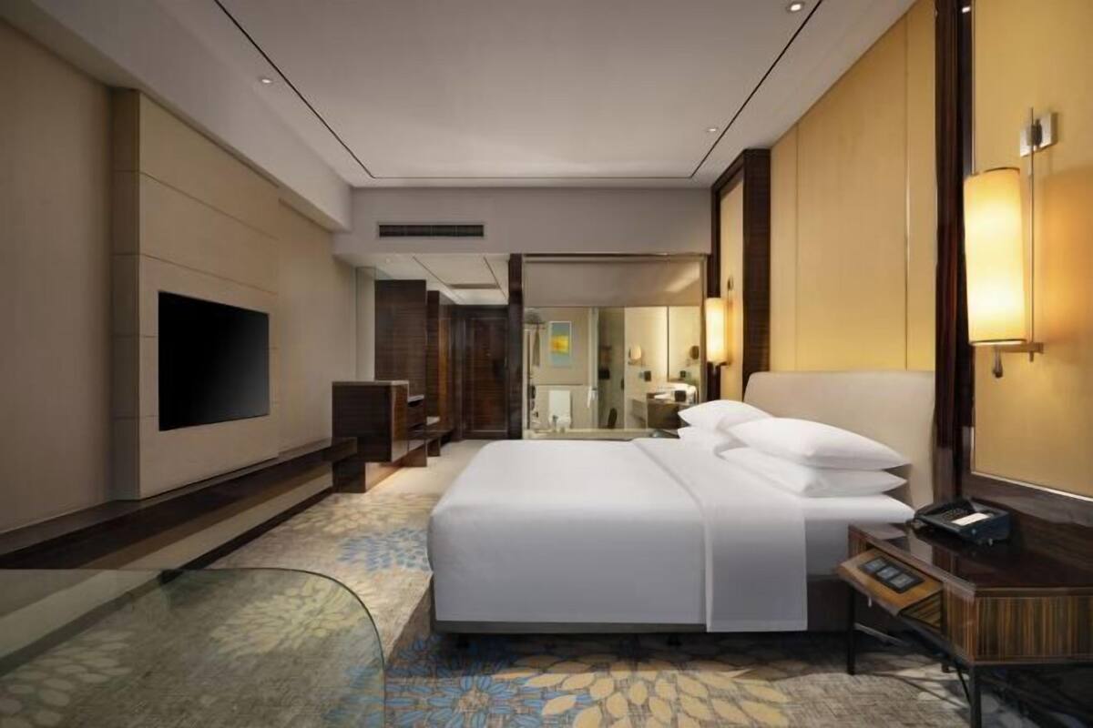 A modern hotel room with a large bed, a flat-screen TV, a desk with a lamp, and a glass coffee table.