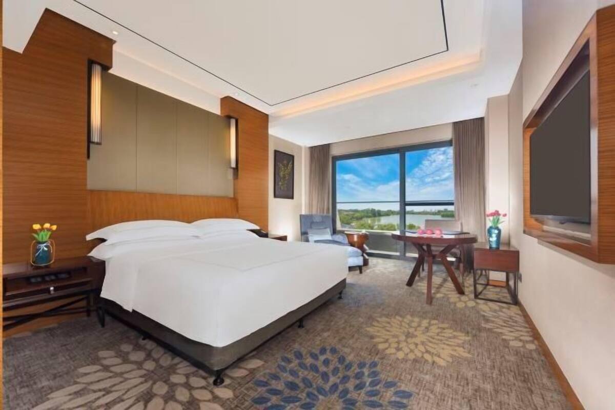 A hotel room with a large bed, a desk, a chair, a TV, and a window with a view.