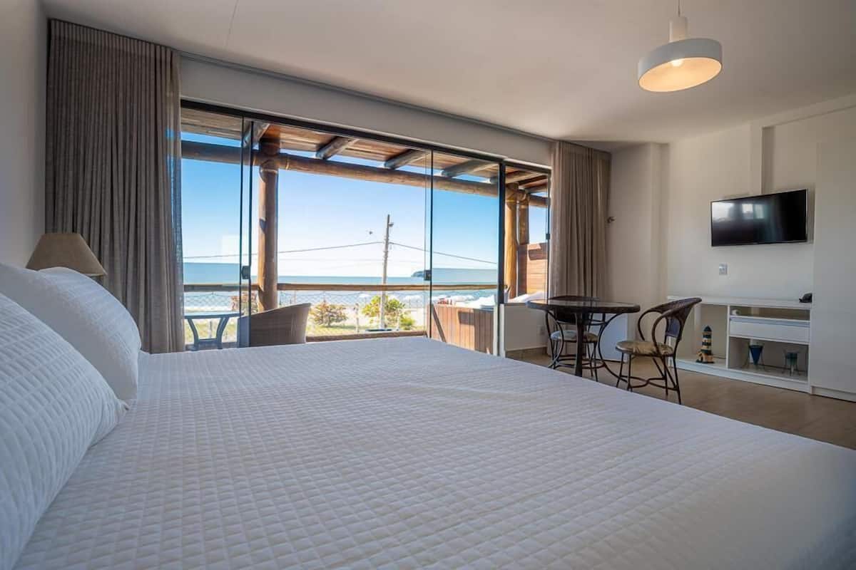 Quarto de hotel com uma cama grande, televisão na parede, varanda com mesa e cadeiras e vista para o mar.
