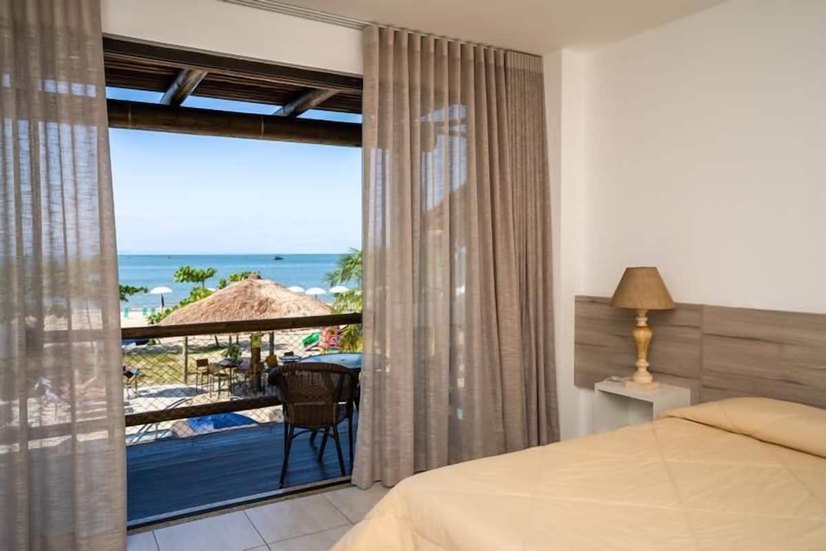 Quarto de hotel com cama, mesa de cabeceira com abajur e vista para a praia e o oceano através de uma grande janela.