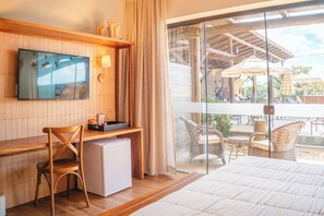 A hotel room with a bed, a desk with a computer, a chair, a small refrigerator, a TV on the wall, and a balcony with outdoor furniture. - Pousada Pedra da Ilha (Penha)