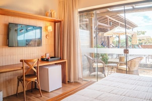 Habitación de hotel con cama, escritorio con computadora, silla, refrigerador pequeño, televisor en la pared y balcón con mobiliario exterior.