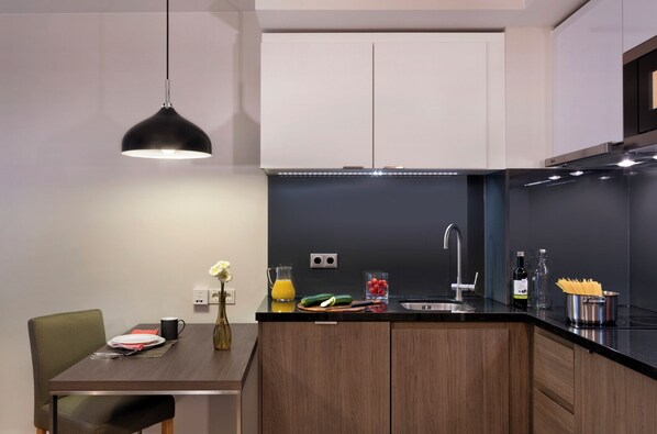 A modern kitchen with a dark countertop, a sink, and a dining area with a chair and table. - Citadines Michel Hamburg (Hamburg)
