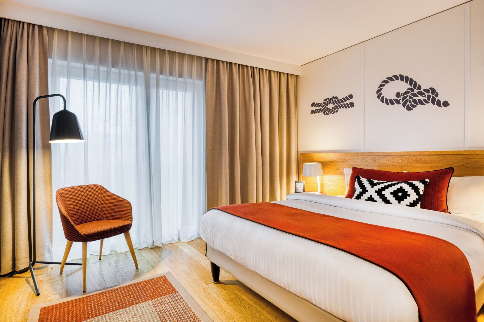 Quarto de hotel com cama, uma cadeira, abajur e janela com cortinas.