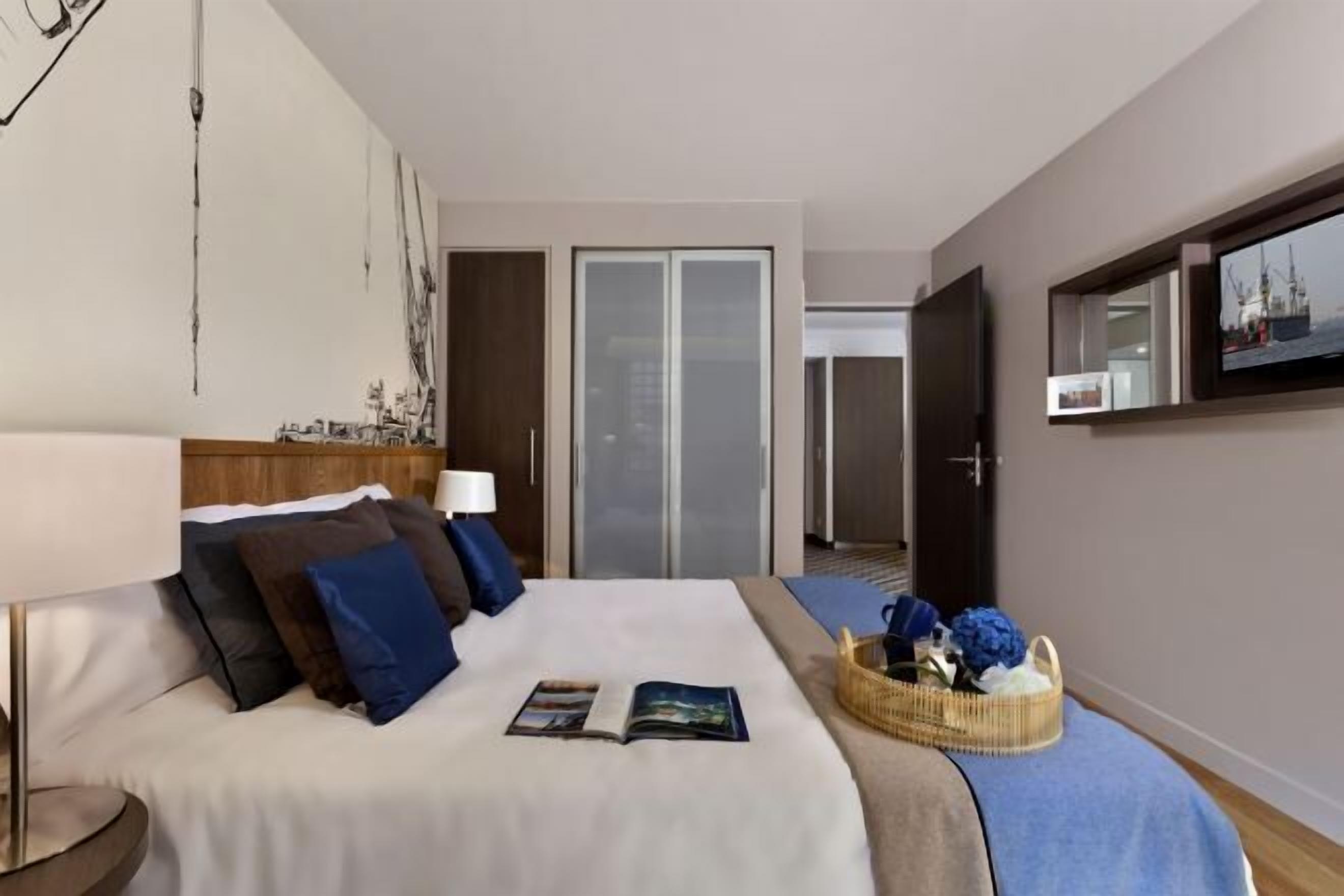 Ein Hotelzimmer mit einem Bett, einem Nachttisch, einer Lampe, einem Buch, einem Korb mit Blumen und einem an der Wand befestigten Fernseher.
