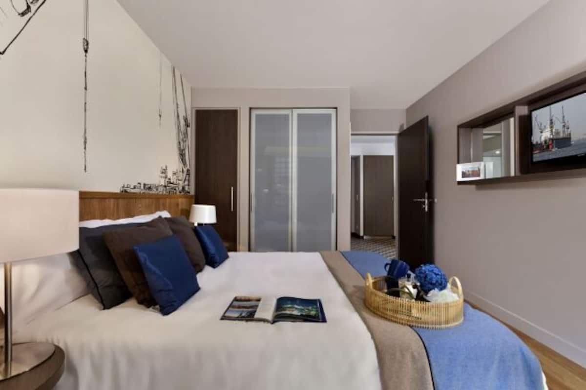 Ein Hotelzimmer mit einem Bett, einem Nachttisch, einer Lampe, einem Buch, einem Korb mit Blumen und einem an der Wand befestigten Fernseher.