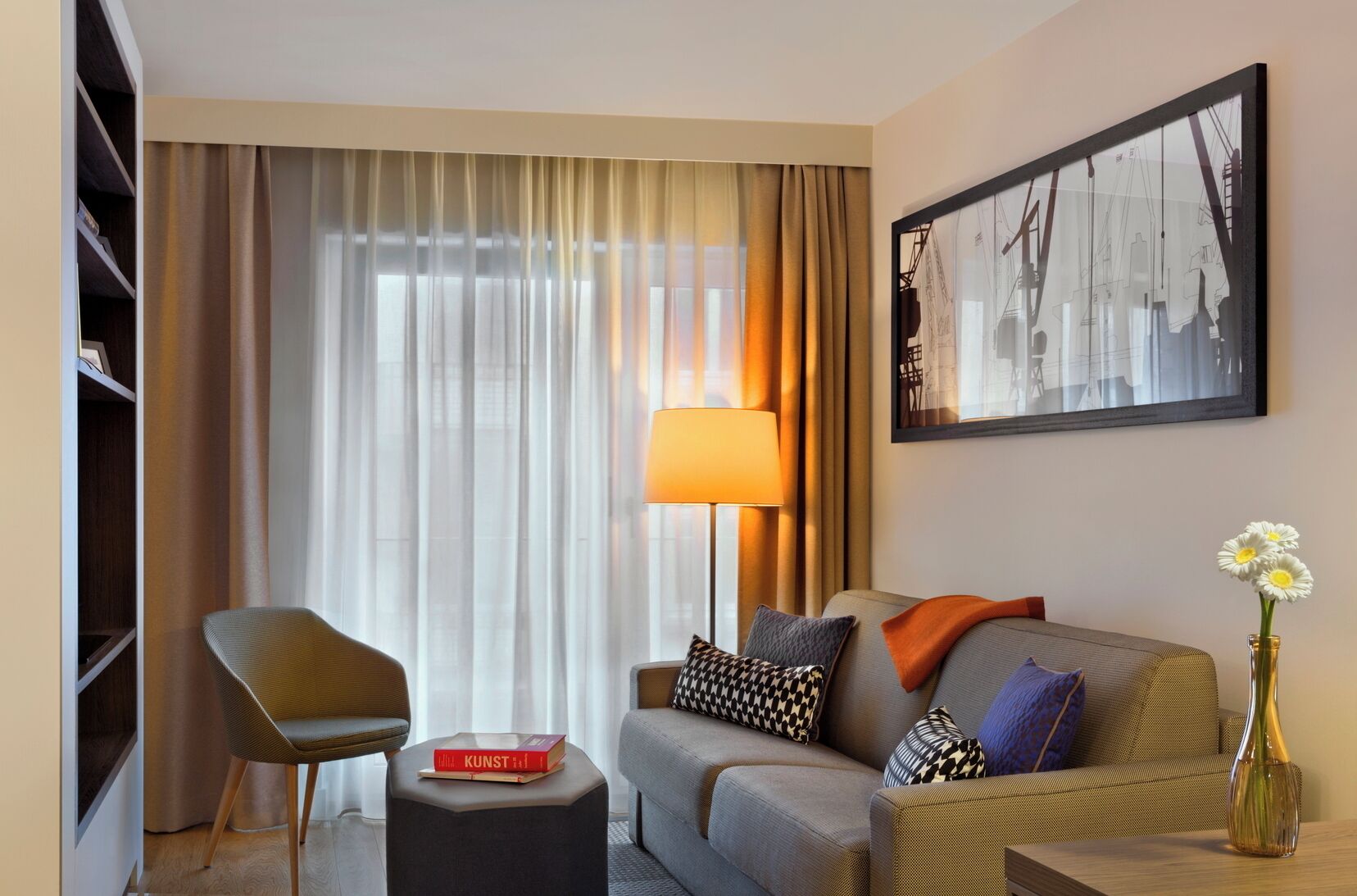 A modern hotel room with a sofa, armchair, and a lamp. There is a bookshelf and a framed picture on the wall.
