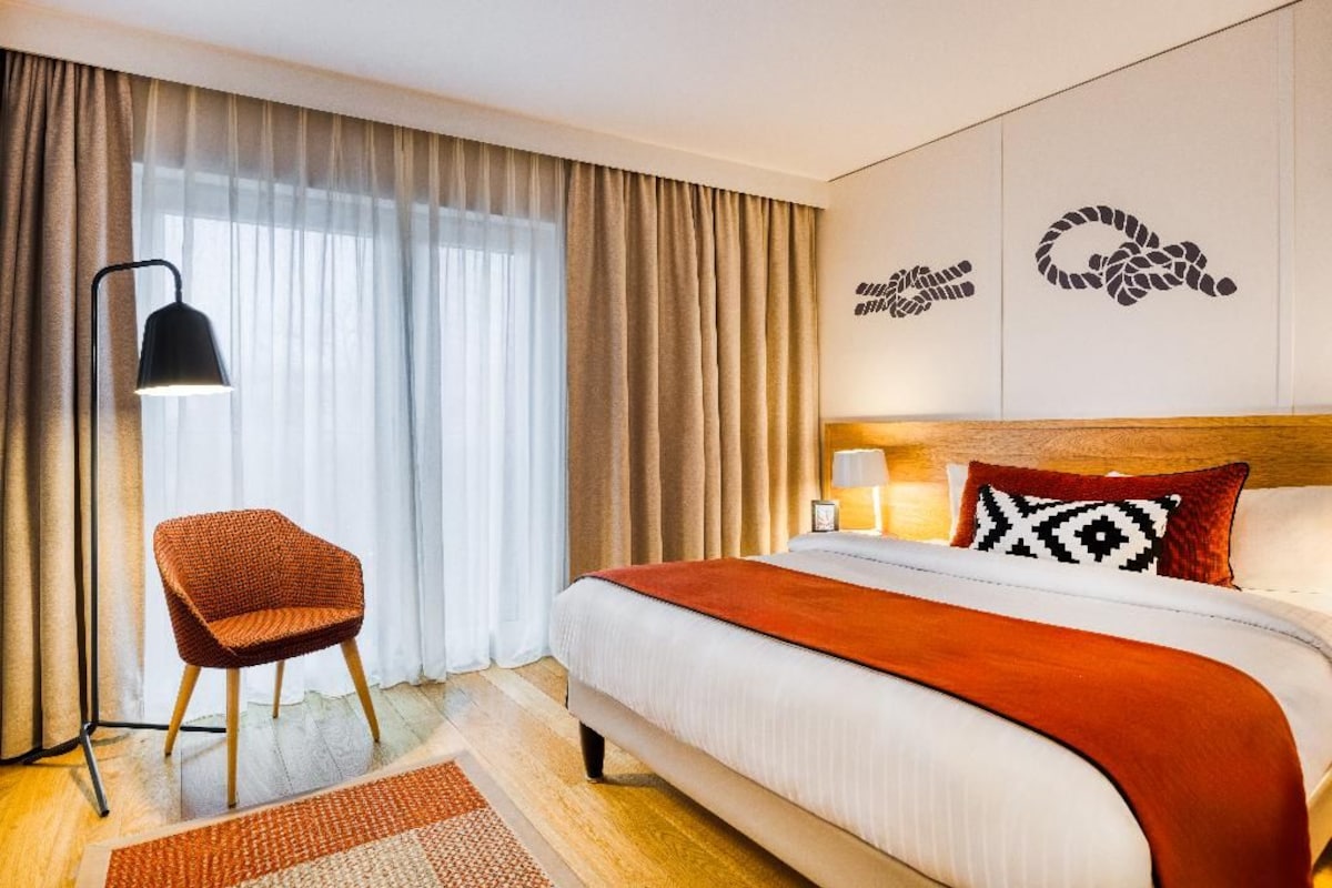 A hotel room with a bed, a chair, a lamp, and a window with curtains.