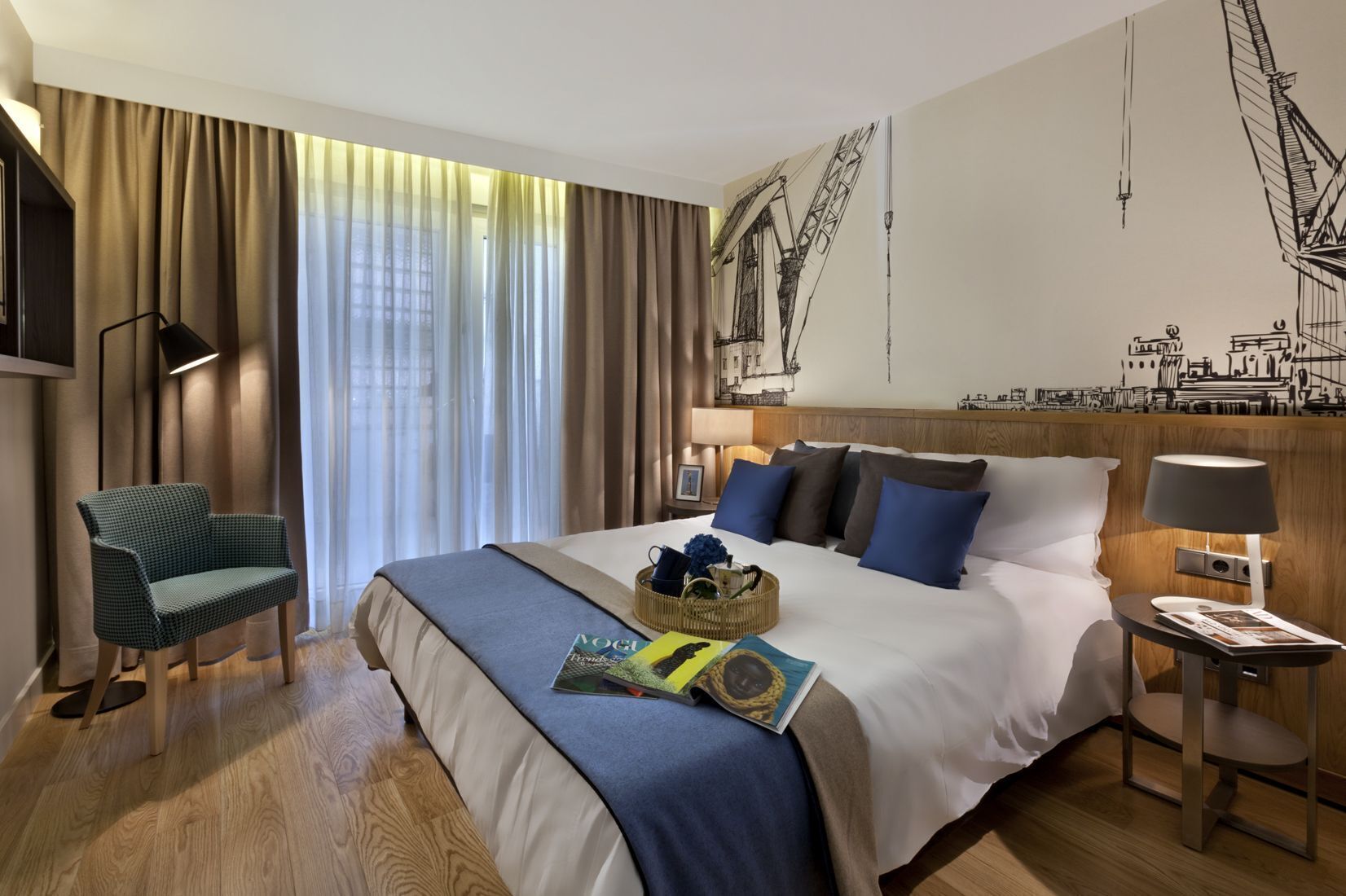 Hotellihuone, jossa on sänky, tuoli, lamppu, seinällä oleva taideteos, jossa on kuvattuna silta, ja ikkuna, jossa on verhot.