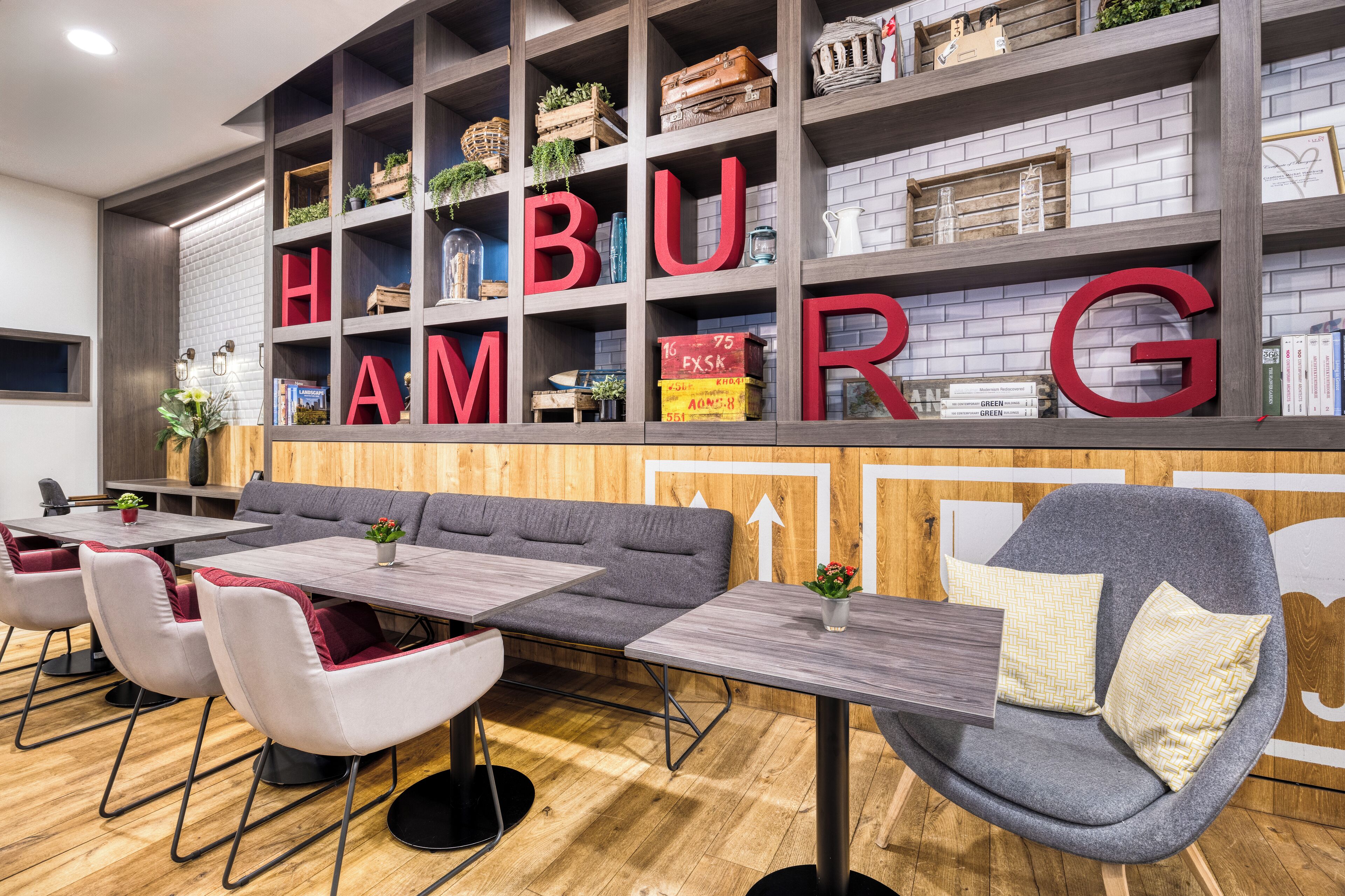 Un intérieur moderne avec une grande bibliothèque où le mot « HAMBOURG » est écrit en lettres rouges, un coin salon avec des canapés et des tables gris, et un plancher en bois.