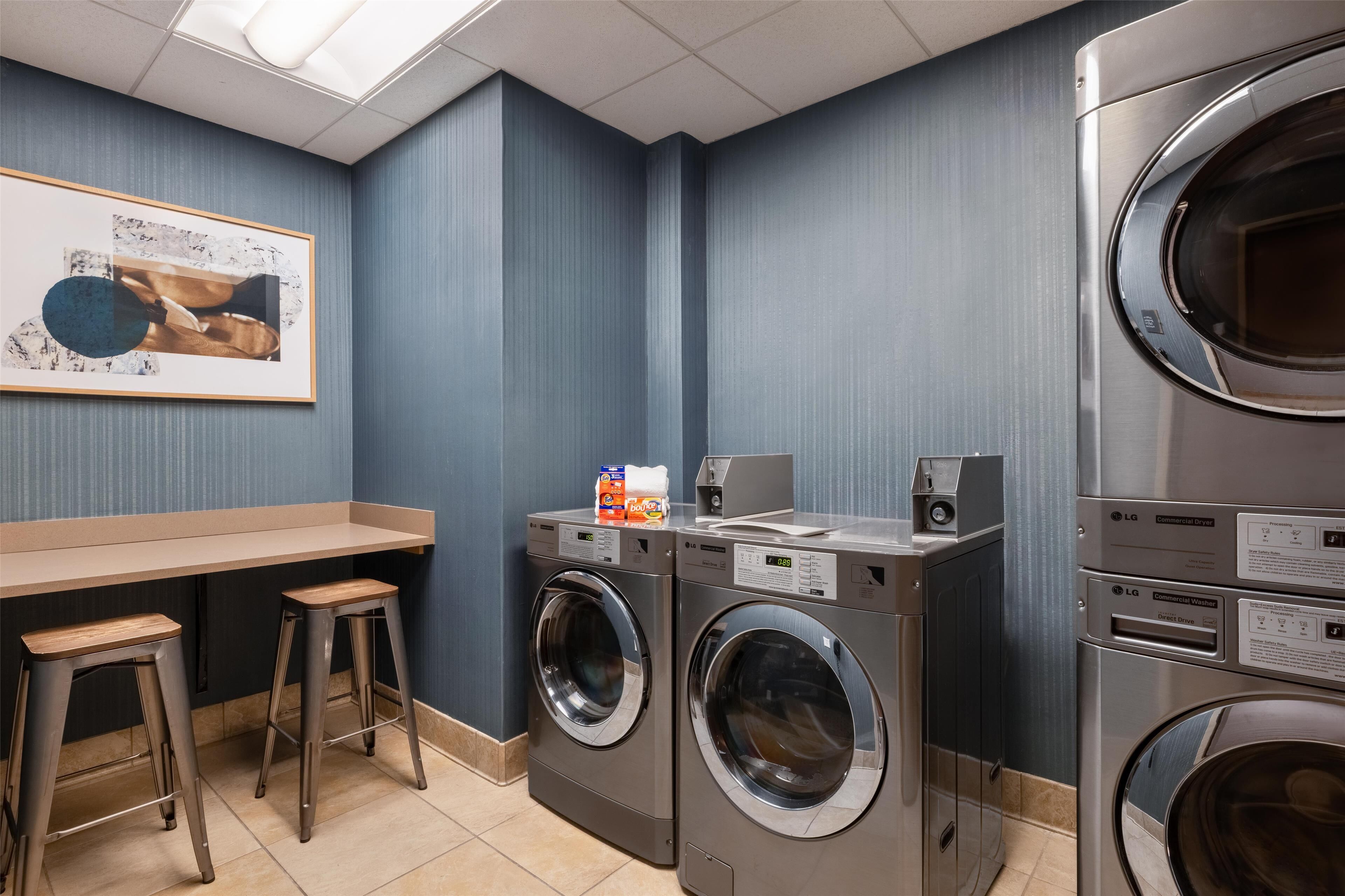 A laundry room with two washing machines, a drying machine, a countertop with a coffee maker, and a framed picture on the wall.