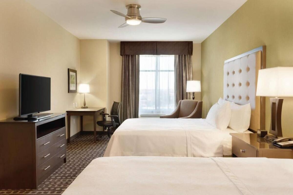 A hotel room with two beds, a desk, a television, a chair, and a ceiling fan.