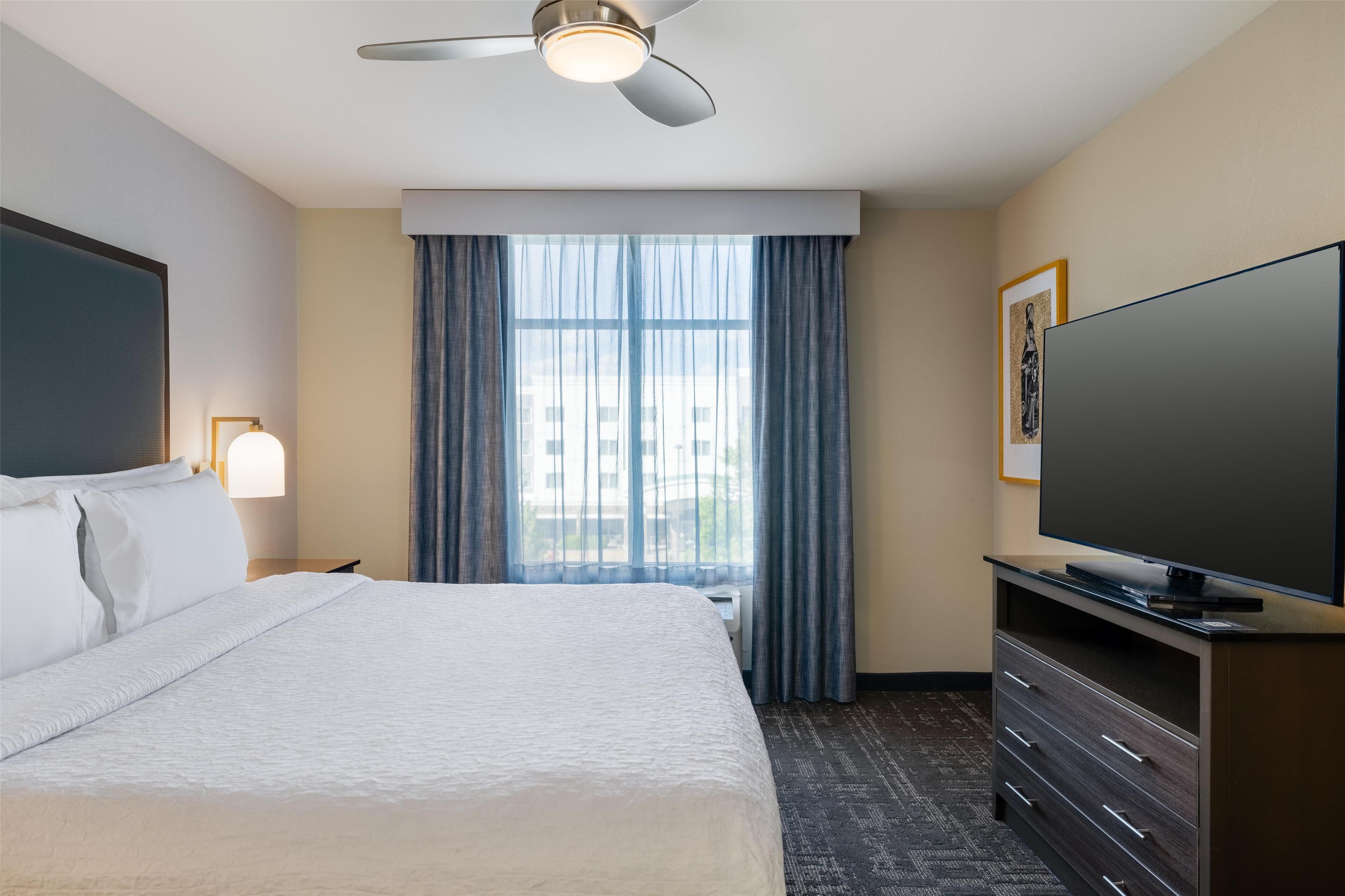 A hotel room with a bed, a flat-screen TV, a ceiling fan, a window with curtains, and a framed picture on the wall.