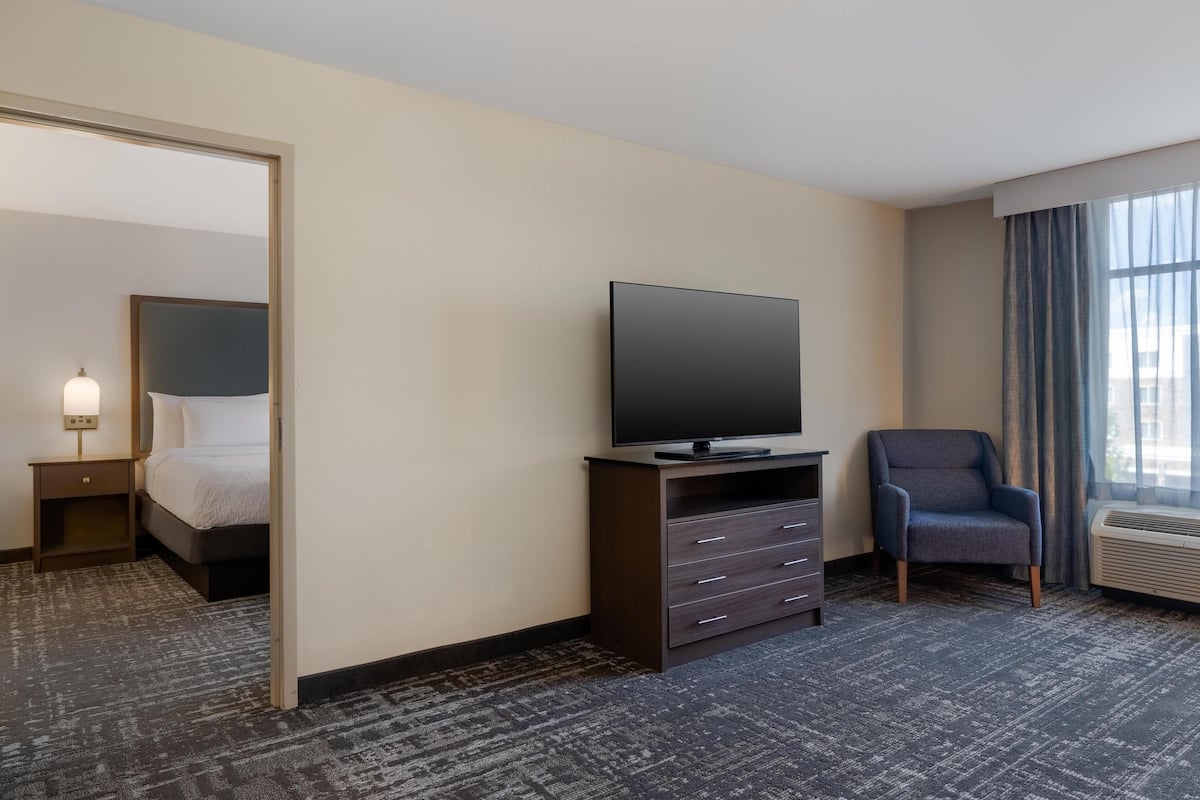 A hotel room with a bed, a television on a stand, a chair, and a large window with curtains.