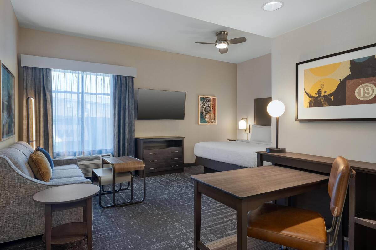 A hotel room with a sofa, a desk, a chair, a bed, a TV, a ceiling fan, and artwork on the walls.