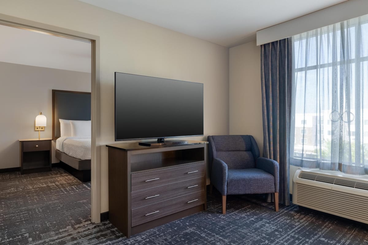 A hotel room with a bed, a flat-screen TV, a chair, a desk, and a window with curtains.