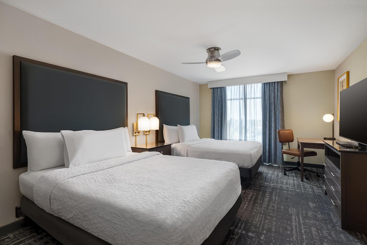 A hotel room with two beds, a desk, a chair, a TV, a ceiling fan, and a window with curtains.