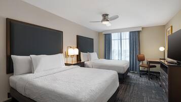 A hotel room with two beds, a desk, a chair, a TV, a ceiling fan, and a window with curtains.