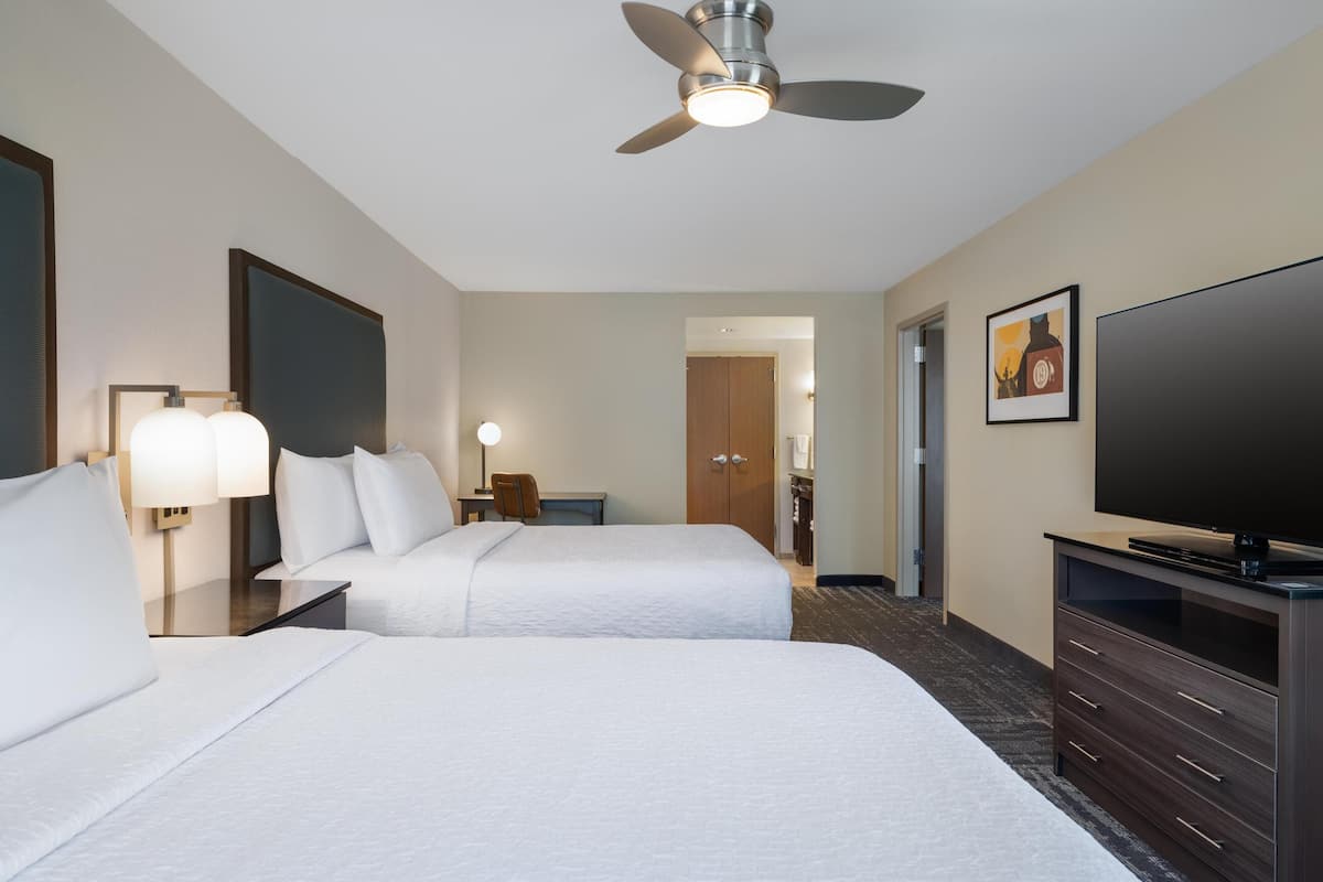 A hotel room with two beds, a ceiling fan, a TV, a desk with a chair, and a door leading to another room.