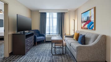 A hotel room with a flat-screen TV, a sofa, a desk with drawers, a standing lamp, and a painting on the wall.