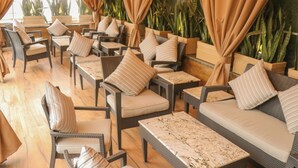 A sunlit indoor seating area with wicker furniture, cushioned chairs, and sofas, featuring a view of green plants and a wooden floor. - Eternity (New Delhi)