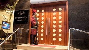 A hotel entrance with a wooden door featuring circular cutouts, a sign reading "ETERNITY Celebrate Forever," and a person in uniform standing on the steps. - Eternity (New Delhi)