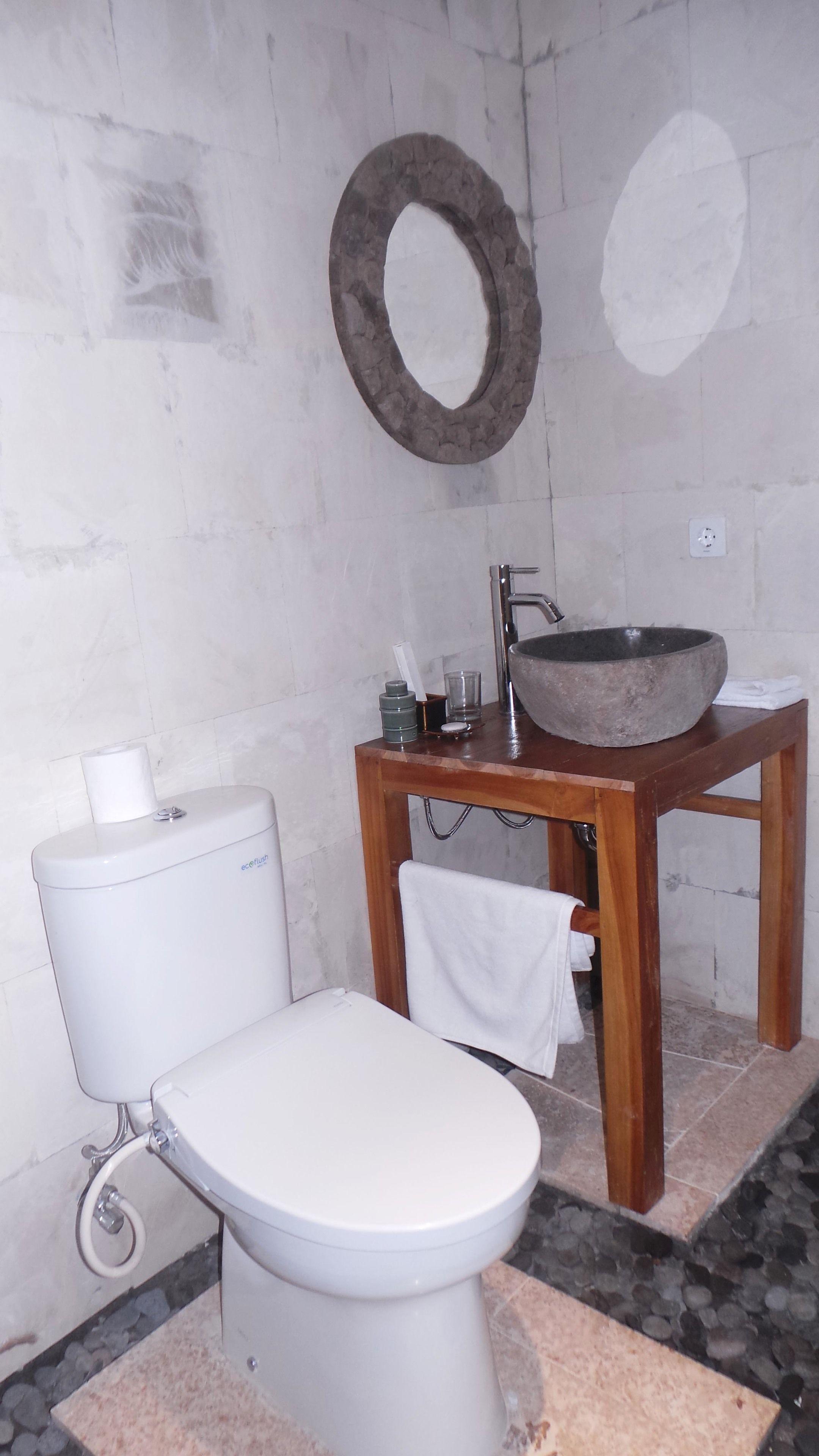Un baño con un inodoro blanco, un tocador de madera con lavamanos de piedra y una toalla colgada en la pared.