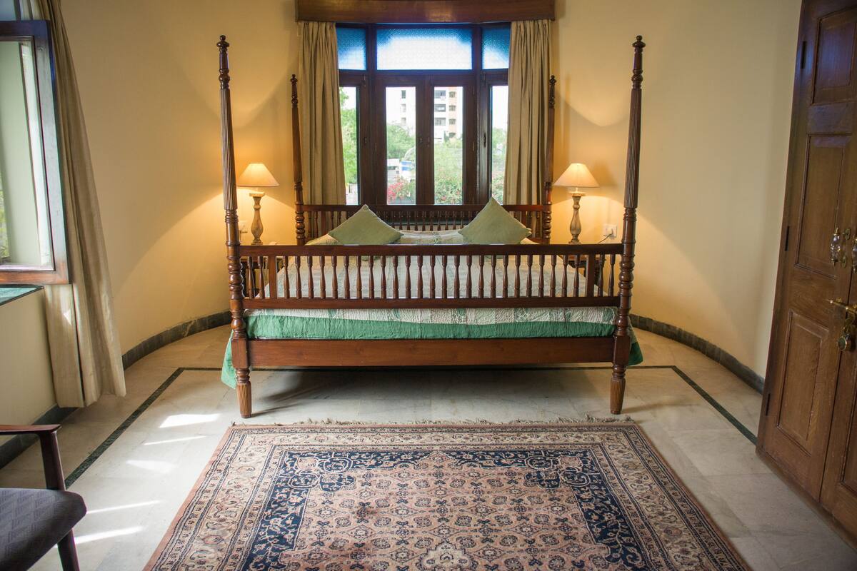 Un lit à baldaquin avec une couvre-lit verte et deux oreillers, flanqué de deux lampes, dans une pièce dotée d’une grande fenêtre et d’un tapis à motifs.