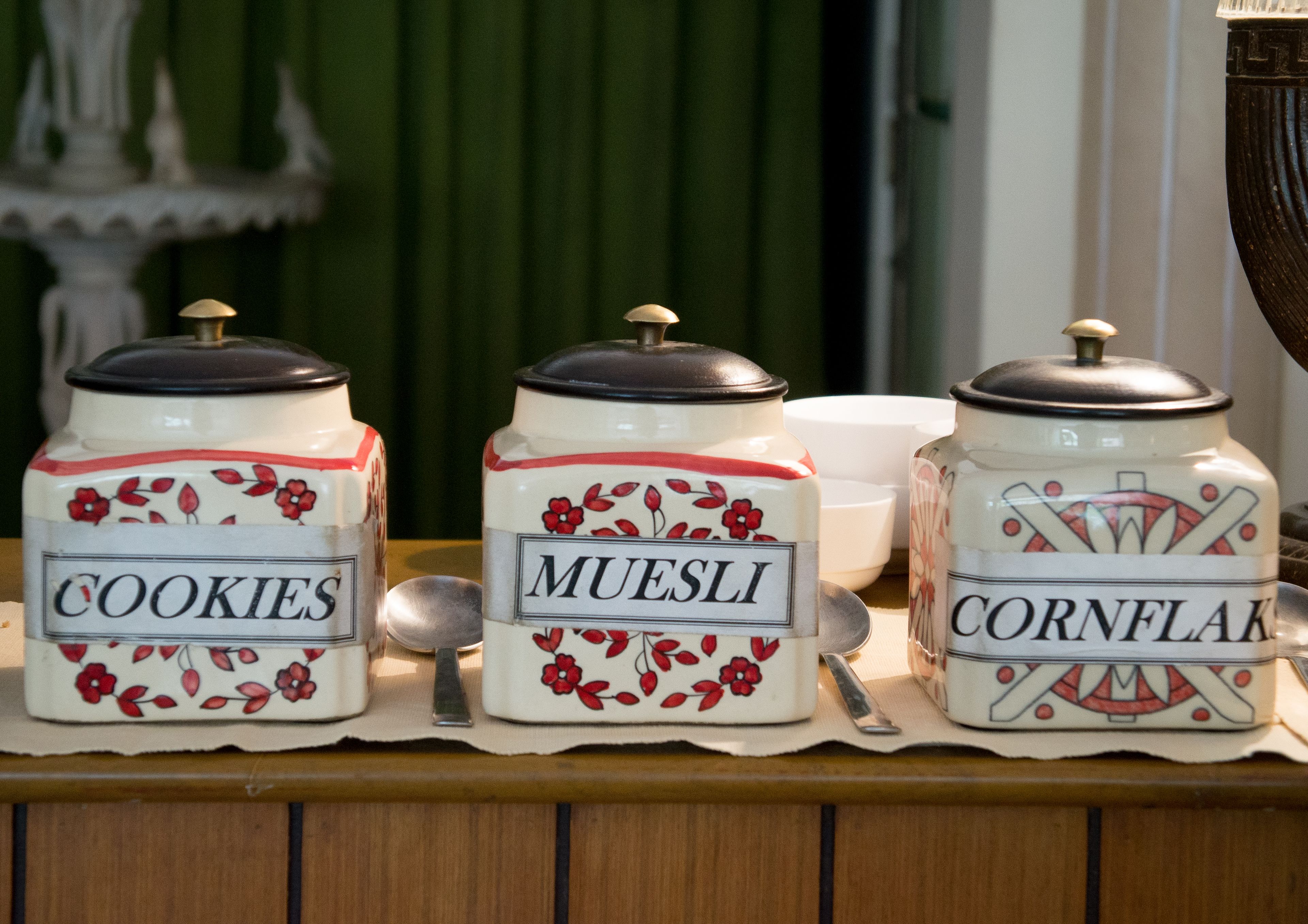 Three ceramic jars labeled 'Cookies,' 'Muesli,' and 'Cornflak' on a wooden table.